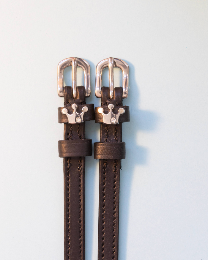 Brown leather ManeJane crown-charm spur straps on a soft blue-grey background