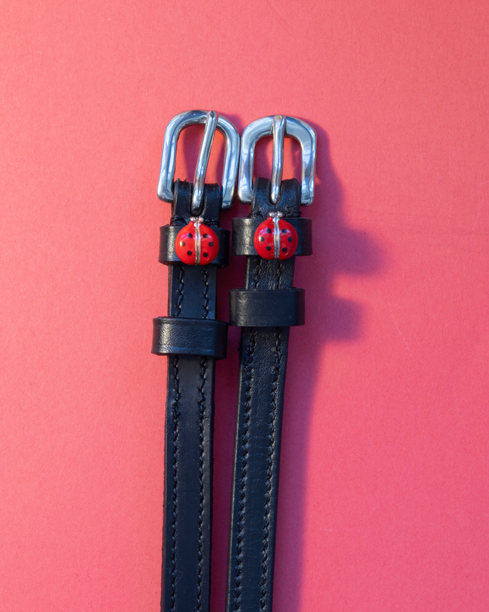 Pair of red ladybug charm spur straps laid flat on a coral background