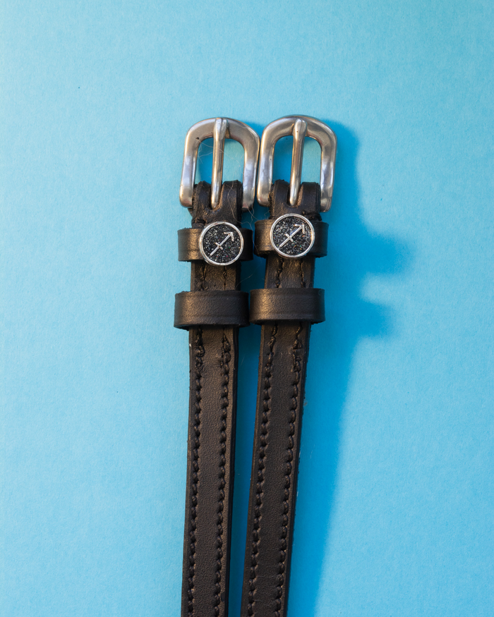 Black leather ManeJane spur straps with round Sagittarius zodiac charms on a blue background