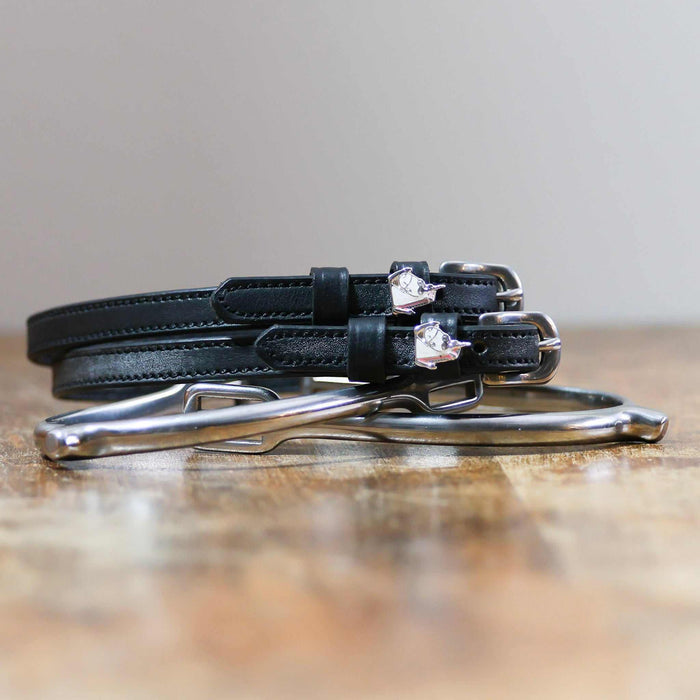 Black unicorn charm spur straps coiled beside silver spurs on a rustic wood surface