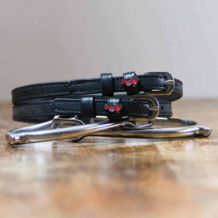 Black leather seahorse spur straps coiled beside a stirrup on a wooden surface