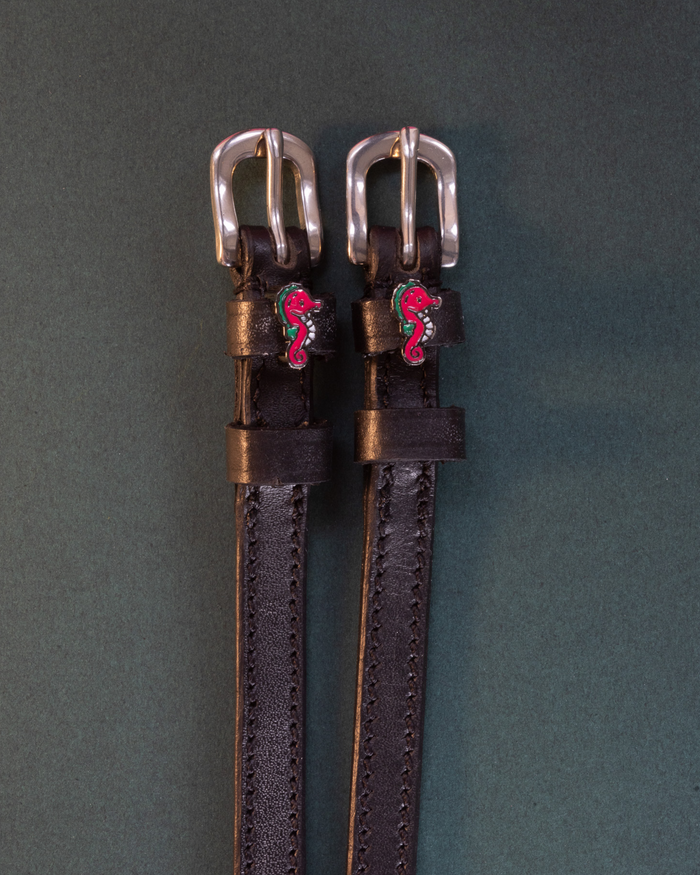Black leather seahorse charm spur straps flat-laid on a dark green background