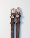 Brown leather ManeJane spur straps with silver shark charms and rose-gold buckles on grey