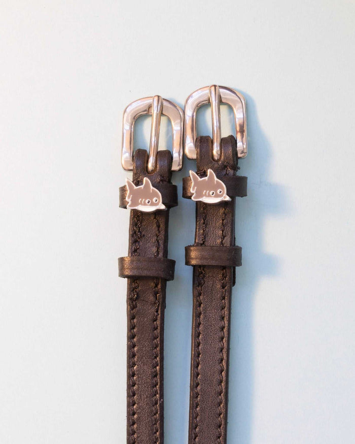 Brown leather ManeJane spur straps with silver shark charms and rose-gold buckles on grey