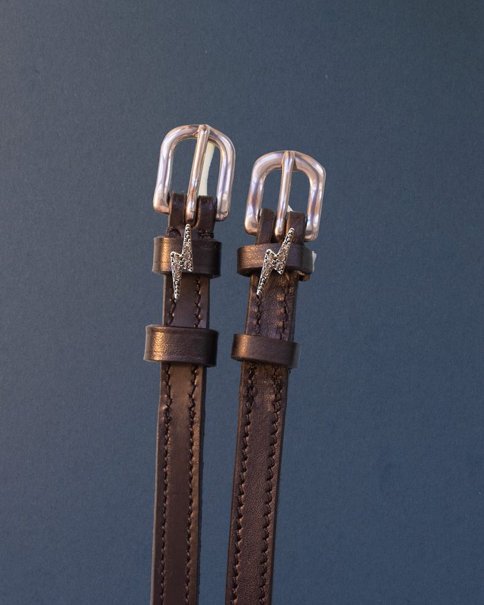 Brown leather ManeJane spur straps with silver rhinestone lightning bolt charms on slate blue