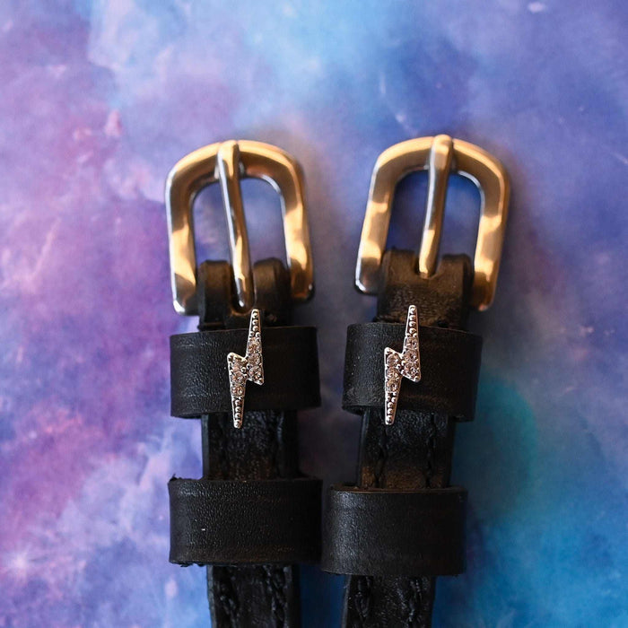 Close-up of rhinestone lightning bolt charms on black spur straps over a galaxy backdrop