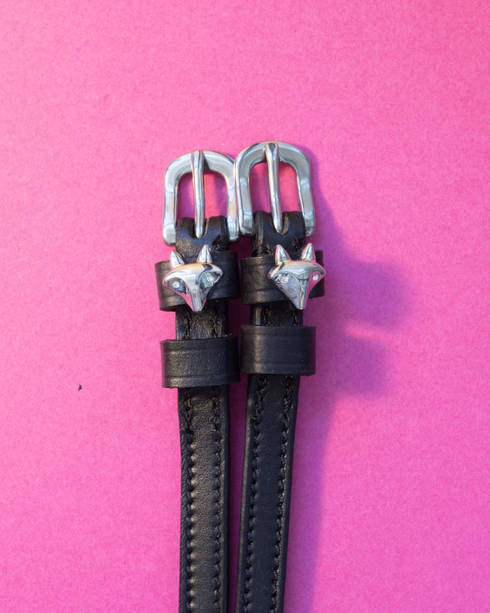 Black leather ManeJane spur straps with silver 3D fox head charms on a pink background