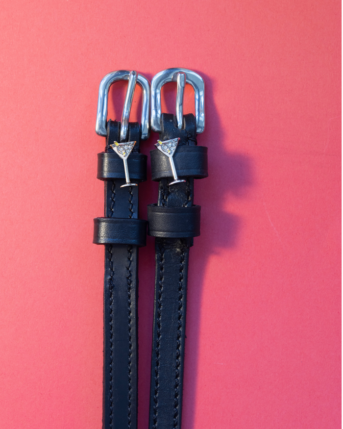 Black leather ManeJane spur straps with silver martini glass charms flat-laid on coral