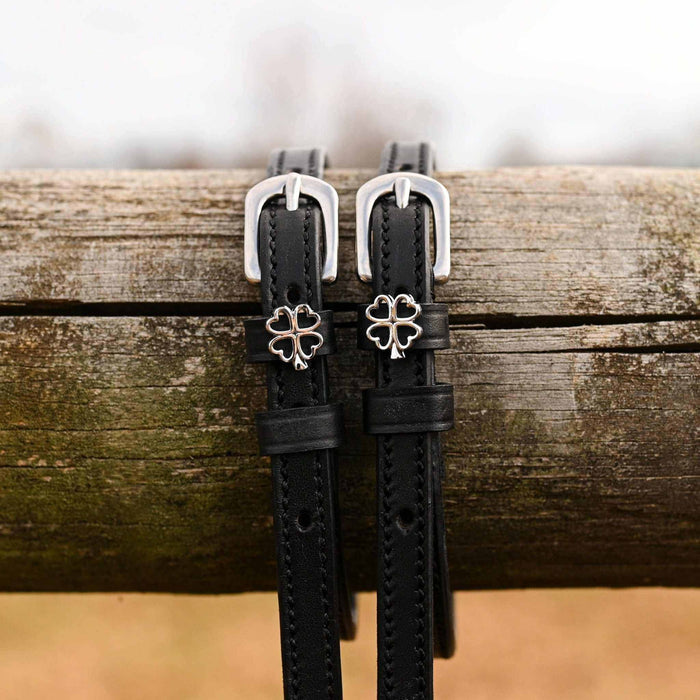 Silver outline clover charms on black leather spur straps draped over a wooden fence rail