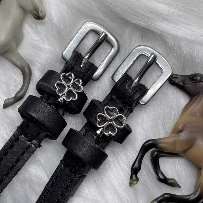 Black leather silver clover spur straps on white fur beside toy horse figurines