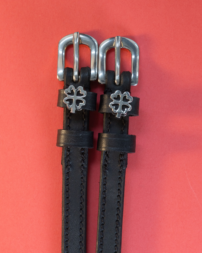 Black leather ManeJane spur straps with silver outline clover charms on a red background