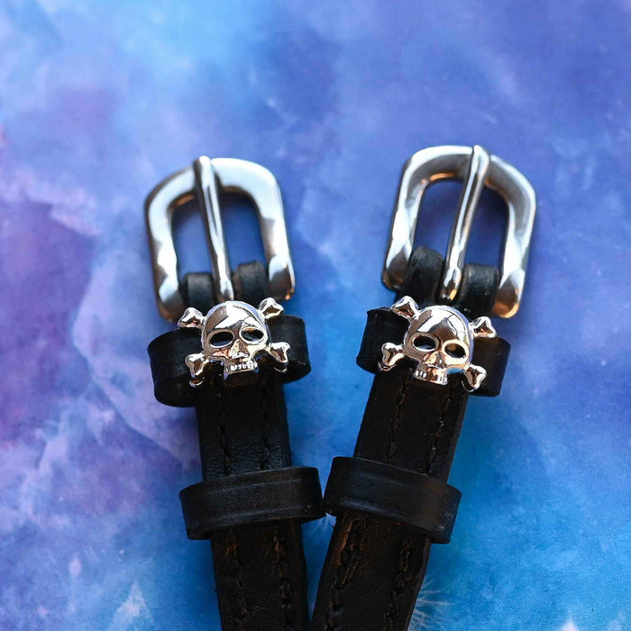 Close-up of silver skull and crossbones charms on spur straps over a purple marble backdrop