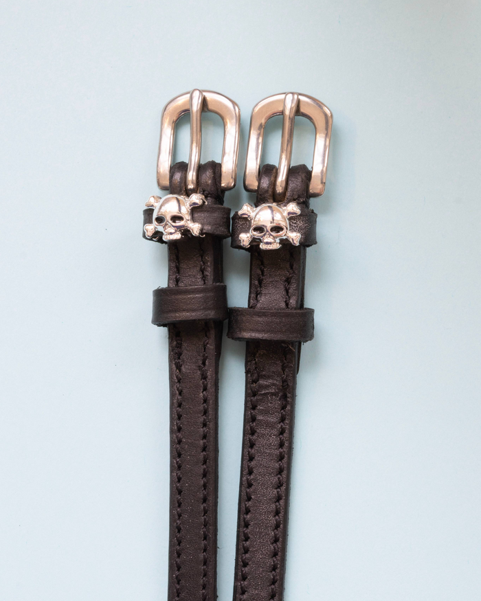 Brown leather skull and crossbones spur straps with rose gold buckles on pale blue