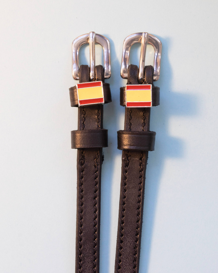 Brown leather ManeJane spur straps with Spanish flag charms on a pale blue background