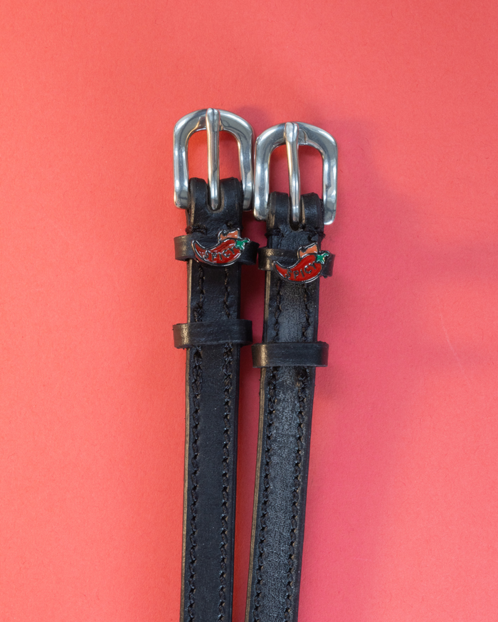 Black leather ManeJane spicy pepper spur straps with red chili charms on a coral background