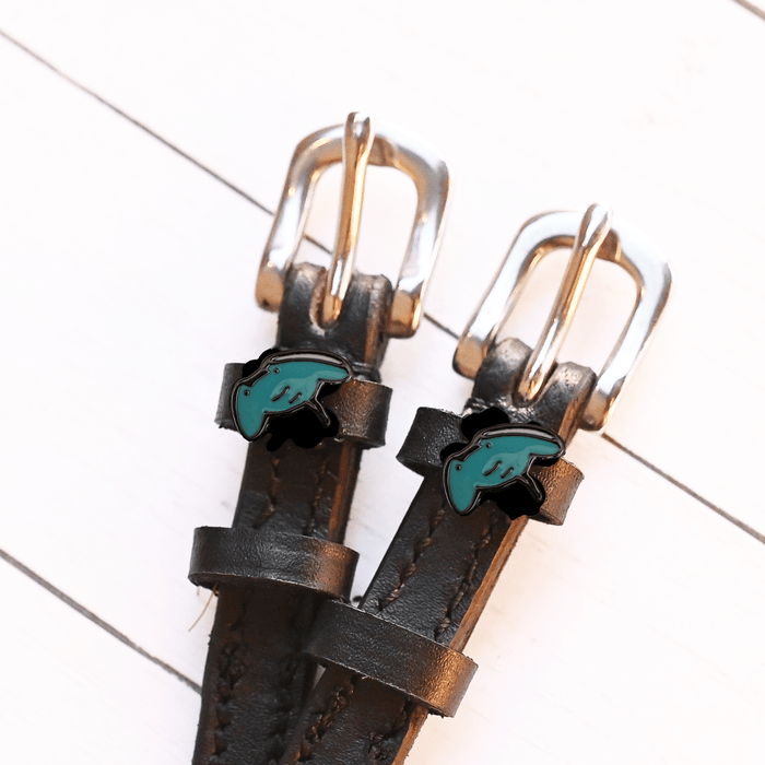Teal stingray charms on black leather spur straps laid on white wood planks, ManeJane
