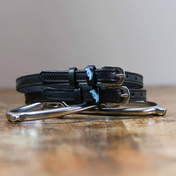 Stacked black stingray spur straps with silver buckles on wood beside a stirrup iron