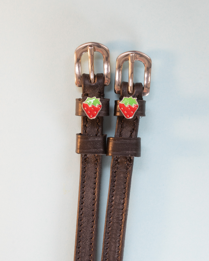 Brown strawberry spur straps with rose-gold buckles and strawberry charms on pale grey, ManeJane
