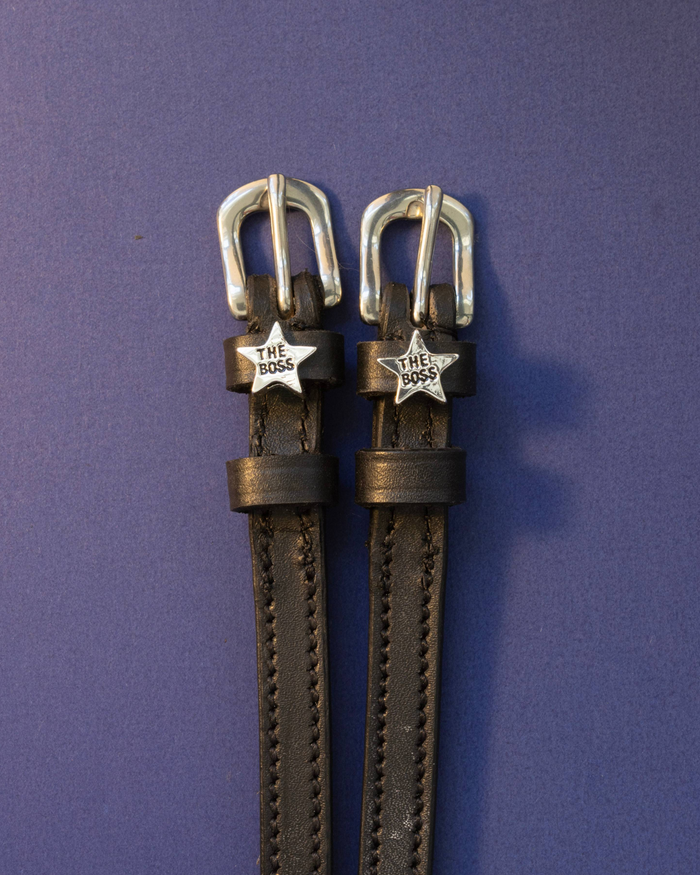 Black leather ManeJane spur straps with silver 'The Boss' star charms on a purple background