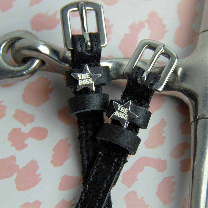 Close-up of silver 'The Boss' star charms on black leather spur straps beside a metal bit