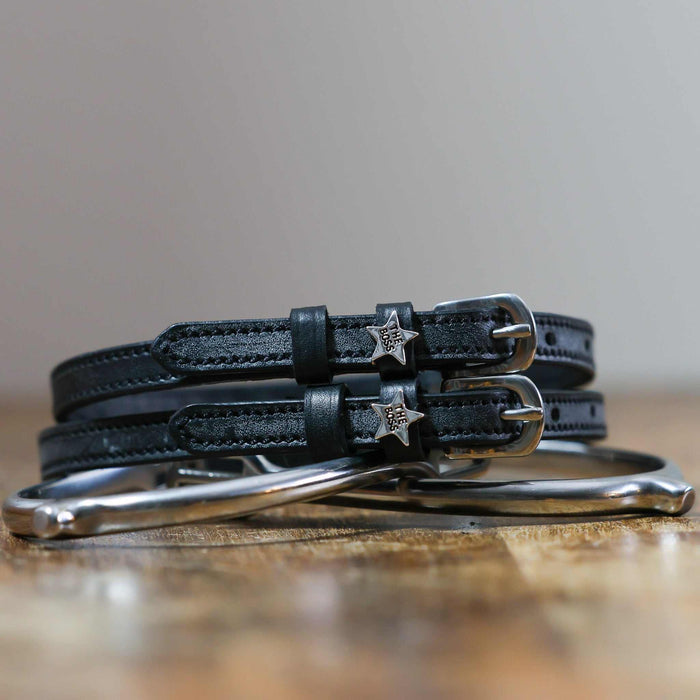 Pair of black leather Boss star spur straps coiled on wood beside a silver bit