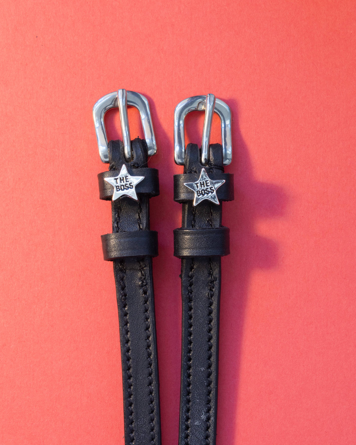 Black leather spur straps with silver 'The Boss' star charms laid flat on a coral background