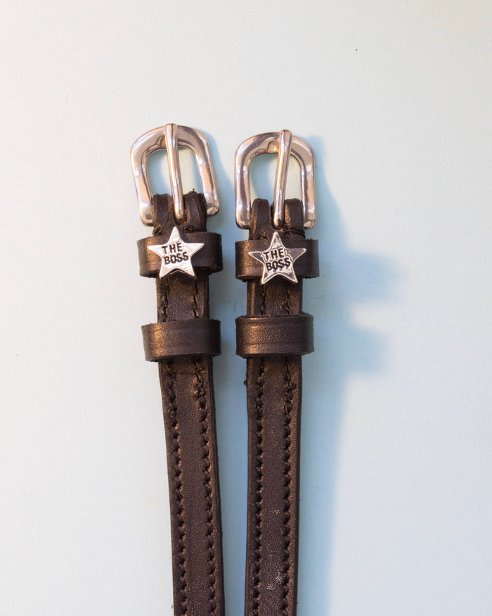 Dark brown leather ManeJane spur straps with silver Boss star charms on a pale background