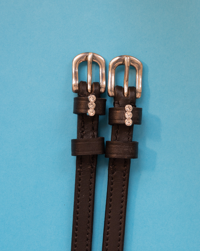 Black leather three-stone charm spur straps with silver buckles on a teal background
