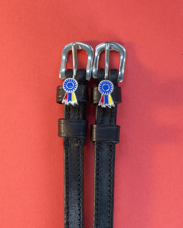 Black leather spur straps with tri-color ribbon rosette charms on a coral background