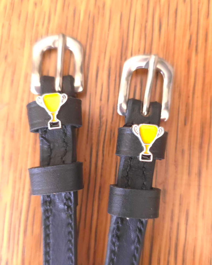 Black leather ManeJane spur straps with yellow trophy cup charms on a wood grain surface