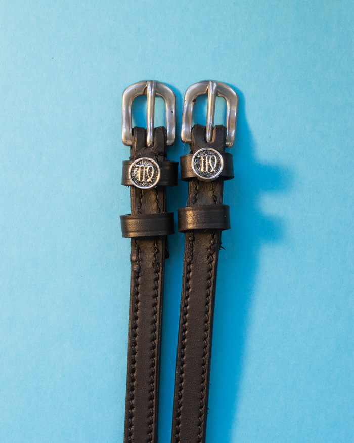 Black leather ManeJane spur straps with round Virgo zodiac charms on a light blue background