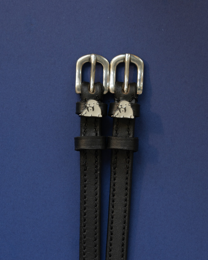Black leather walrus spur straps coiled beside a silver stirrup on a wooden surface