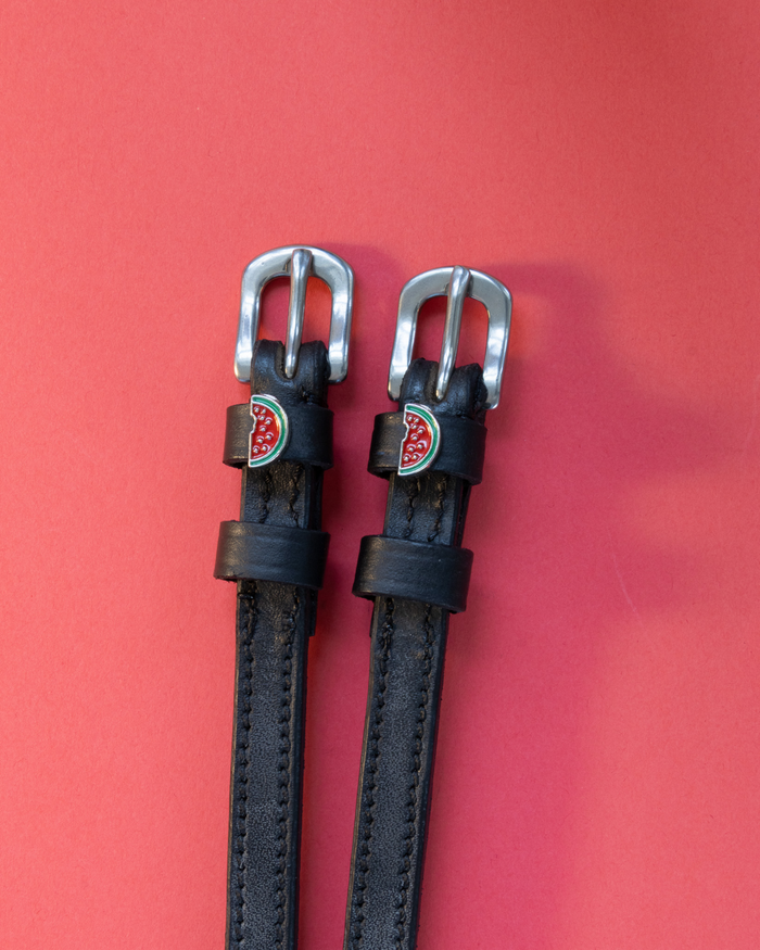 Black leather ManeJane spur straps with silver buckles and enamel watermelon charms on red background
