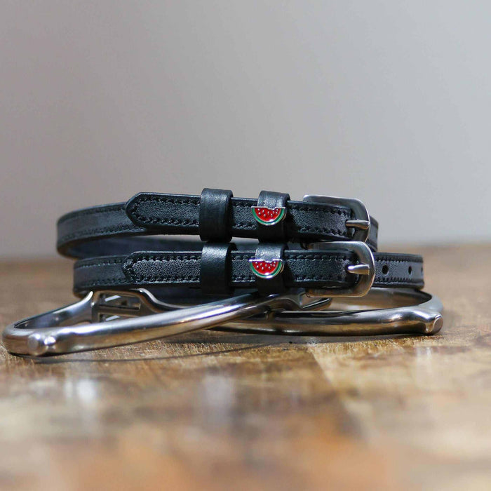 Black watermelon charm spur straps coiled beside a silver spur on a rustic wood surface