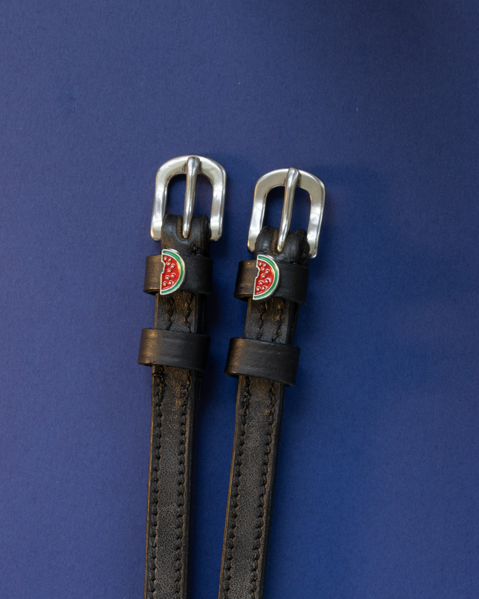 Close-up of black watermelon charm spur straps resting on a silver spur, white background