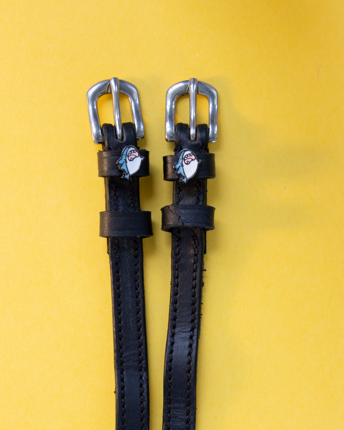 Pair of black leather shark charm spur straps with silver buckles on a yellow background